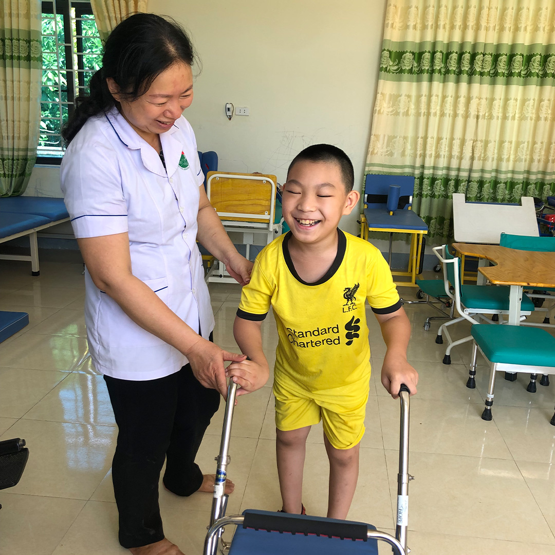 A smiling boy in a yellow team jersey walks with his walker as his therapist cheers him on
