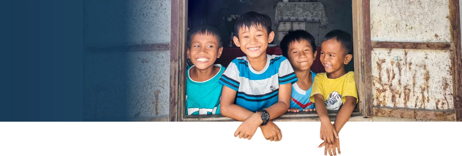 Four smiling boys lean out of a window, their hands hanging over the sill.